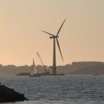A large wind turbine assembled beside a fjord under a wide sky
